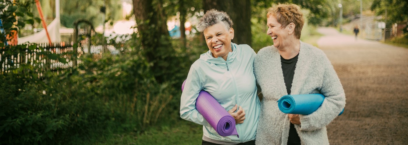 two ladies walking through the park carrying yoga mats and laughing