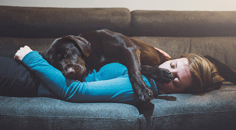 Woman asleep on sofa with a dog laying on top of her.