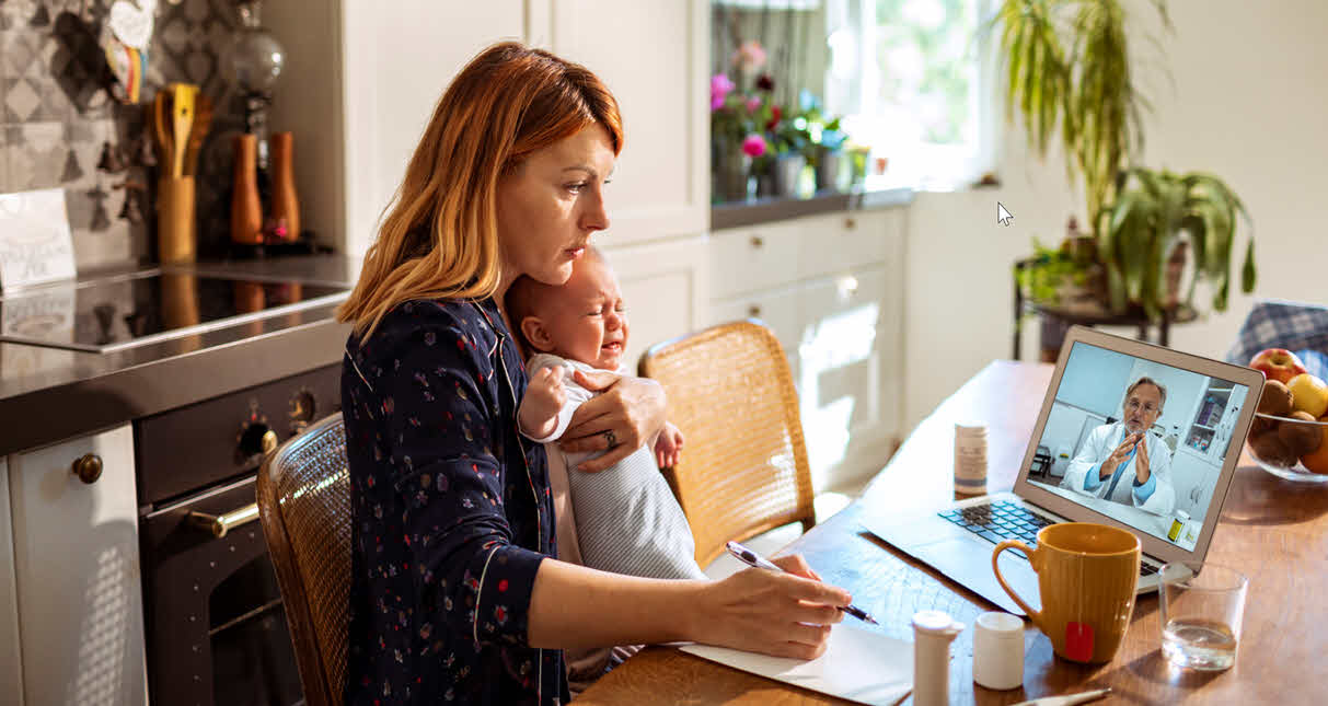 A young mother sat at her kitchen table with her baby. She is speaking to her GP remotely on her laptop.