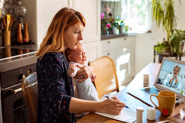 A young mother sat at her kitchen table with her baby. She is speaking to her GP remotely on her laptop.