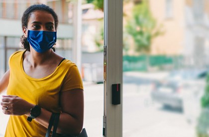 A woman is walking into a shop. She's wearing a blue face mask and a yellow top.
