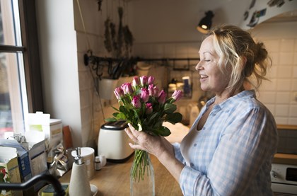 A blonde, smiling middle-aged woman is in her kitchen, arranging fresh flowers.
