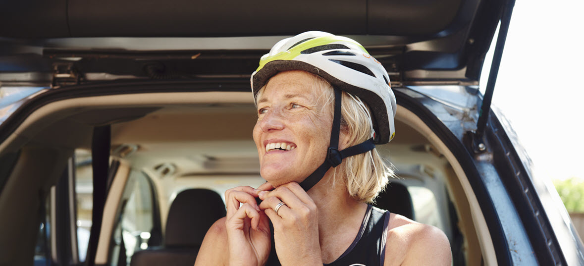 Middle aged woman with short blonde hair in front of her car fastening her cycle helmet whilst smiling 