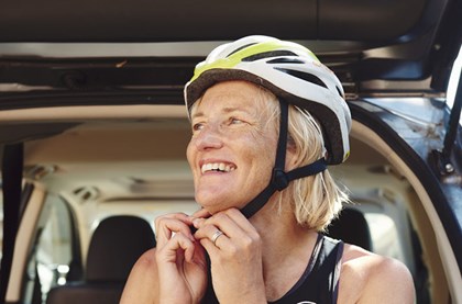 Middle aged woman with short blonde hair in front of her car fastening her cycle helmet whilst smiling 