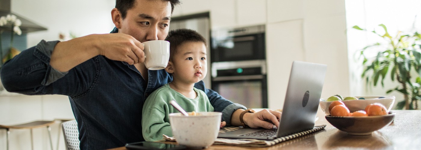 A father working on a laptop, drinking a cup of tea, with a toddler on his lap. 