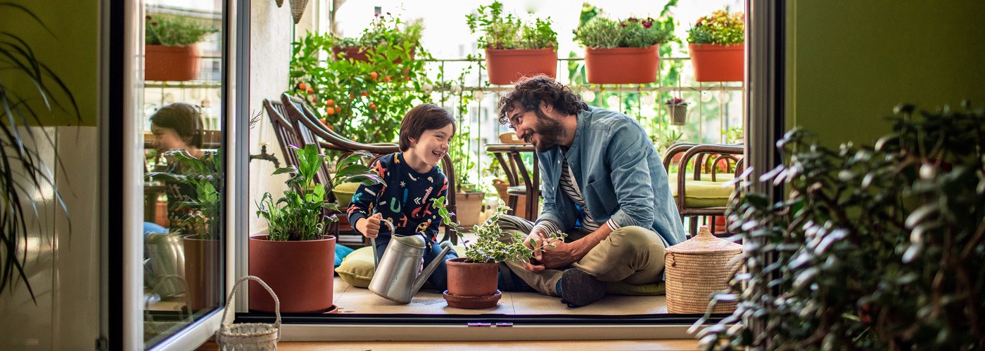 Young man and his son sat on a balcony smiling at one another, surrounded by plants 