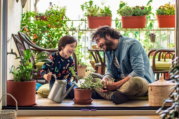 Young man and his son sat on a balcony smiling at one another, surrounded by plants 