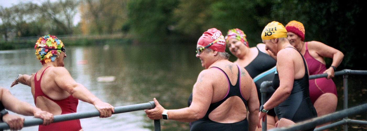A group of middle-aged women in swimming costumes, stood next to a lake 