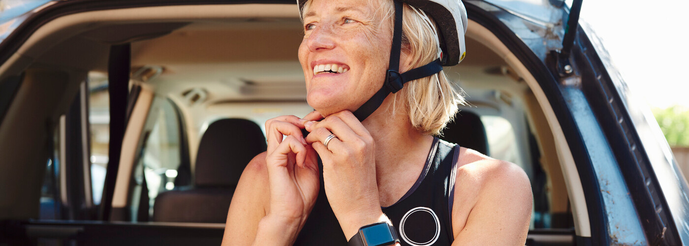 A woman in her 30's with long brown hair with her arm around a man, both smiling with the beach in the background. 