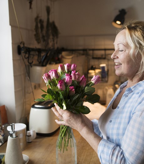 A blonde, smiling middle-aged woman is in her kitchen, arranging fresh flowers.