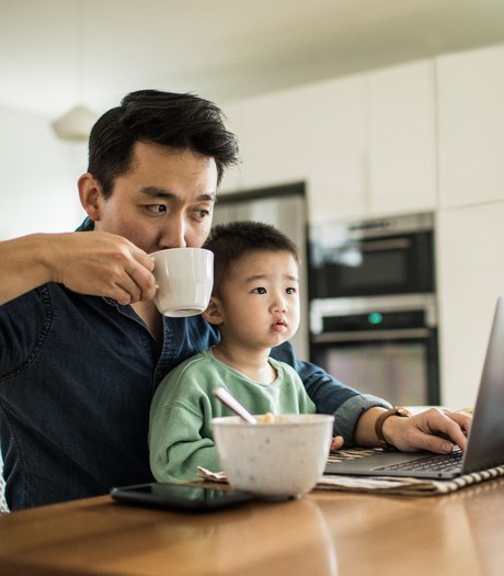 A father working on a laptop, drinking a cup of tea, with a toddler on his lap. 