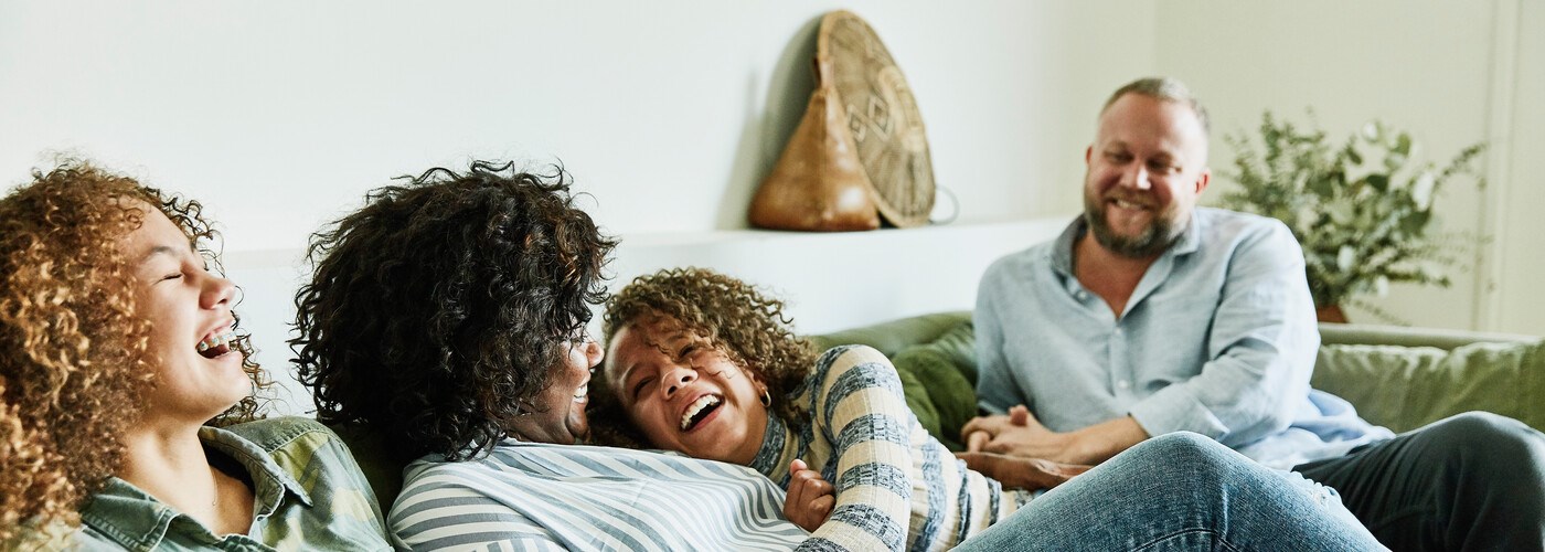 Laughing daughter embracing mother while sitting on couch with family in living room
